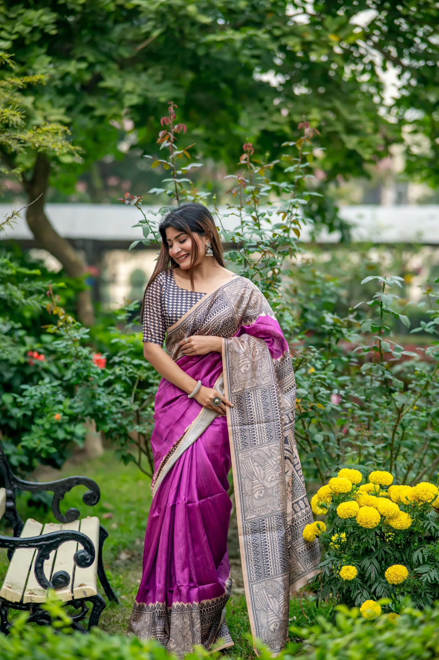 Woman in a deep purple saree with Madhubani print standing near a pond with lotus flowers in a lush garden setting