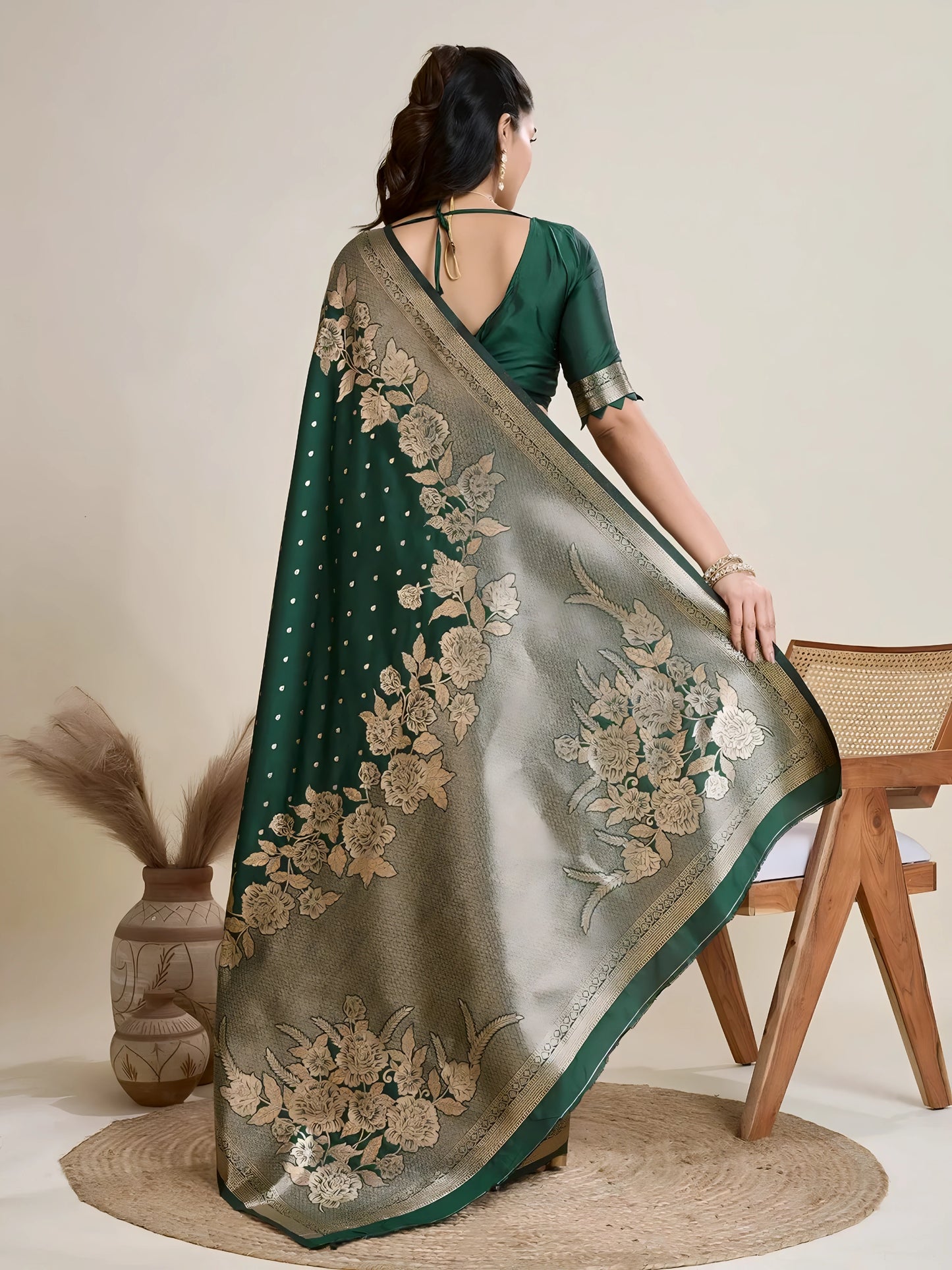 Woman wearing a green Banarasi silk saree with intricate gold floral zari border and pallu, paired with a matching blouse and traditional jewelry, elegantly posing beside a wooden chair in a minimalist studio with neutral decor, ceramic vases, and pampas grass.
