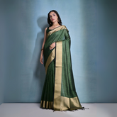 Woman wearing a green silk saree with gold border and traditional jewelry, posing against a plain blue background