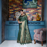 Woman wearing an elegant green silk saree with golden zari floral motifs and a rich gold border, accessorized with traditional gold jewelry, gracefully posing in a stylish vintage-inspired room with a blue cabinet, multicolored brick wall, decorative art, and a patterned armchair.