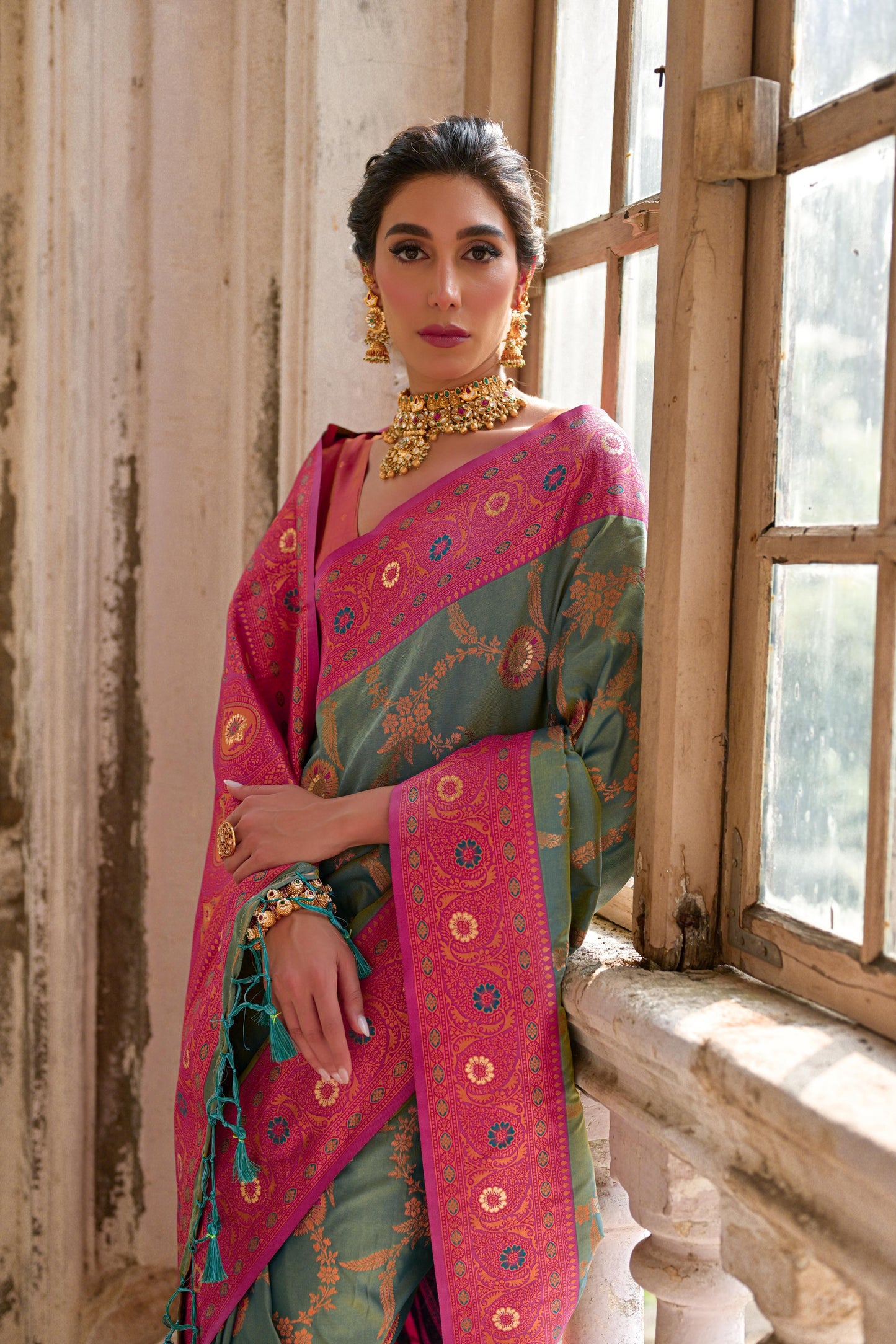 Woman wearing a green silk saree with intricate pink floral zari border and circular motifs, paired with a matching pink blouse and traditional gold jewelry, standing gracefully in a vintage room with large antique wooden windows and ornate architectural details.