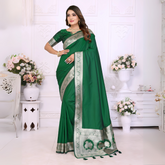 Woman wearing an elegant emerald green silk saree with a rich silver zari border and matching blouse, posing gracefully in a white floral-themed room with classic wall panels and decorative flower arrangements.