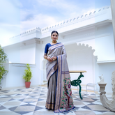 Woman wearing a grey silk saree with blue border and blouse, featuring a handpainted pallu with tree and bird design, posing in a white heritage courtyard with patterned tile flooring and potted plants.