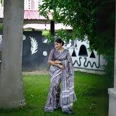 Woman wearing a grey handloom cotton saree with white tribal-inspired prints and a matching patterned blouse, accessorized with silver jewelry, standing on green grass in front of a wall with tribal art motifs in an outdoor garden setting.