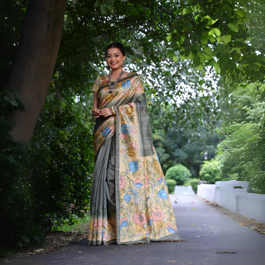 Woman wearing a grey saree with vibrant Madhubani-inspired floral print border and pallu, paired with a matching blouse, standing gracefully on a garden pathway surrounded by lush green trees and foliage.