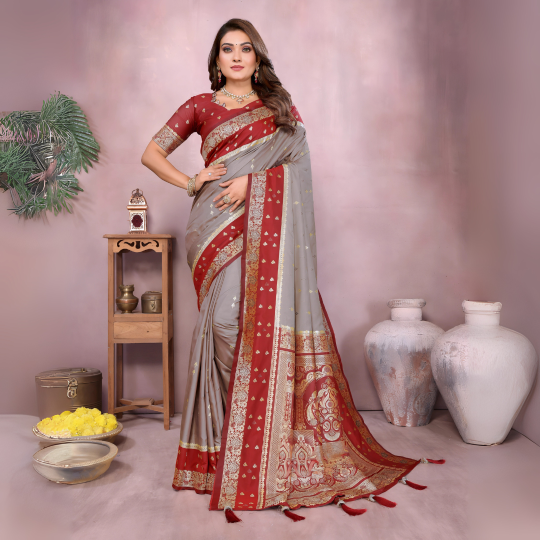 Woman draped in an elegant grey Banarasi saree featuring a rich red and gold embroidered border with intricate traditional motifs, paired with a matching red blouse. She poses gracefully in a modern studio decorated with rustic clay pots, a wooden side table holding brass decor, yellow flowers in a bowl, and a small green branch adding a natural accent to the setting.Woman draped in an elegant grey Banarasi saree featuring a rich red and gold embroidered border with intricate traditional motifs, paired with