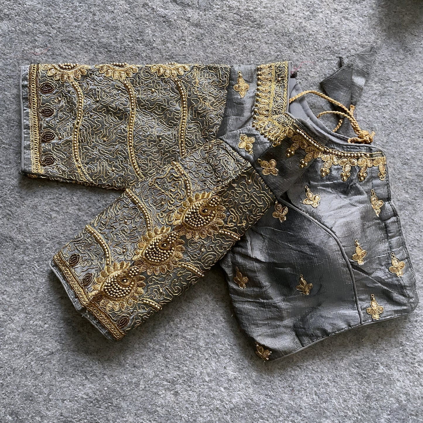 Traditional grey silk blouse featuring intricate gold embroidery and beadwork with floral and paisley motifs on the sleeves and neckline, displayed flat on a textured surface with a bunch of artificial flowers and a woven mat in the background.