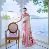 Woman wearing an ivory silk saree with pink border and handpainted floral pallu, styled with gold jewelry, posing in a traditional arched pavilion overlooking a serene lake and distant mountains.