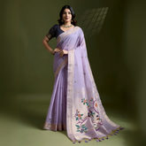 Woman wearing a lavender silk saree with subtle embroidered motifs and a cream-colored border, featuring multicolor floral designs on the pallu, paired with a contrasting black blouse and traditional jewelry, elegantly posing in a studio with olive green walls and a window-patterned light effect.