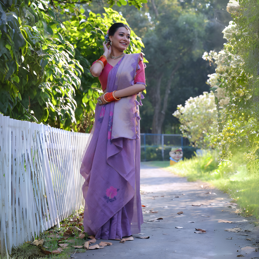 A woman models an elegant Lavender Purple Soft Muga Cotton Saree. The pallu features subtle Resham floral weaving in pink and purple. She wears a contrasting bright pink blouse and is accessorized with a pearl choker necklace and vibrant orange bangles, posing outdoors by a white fence.