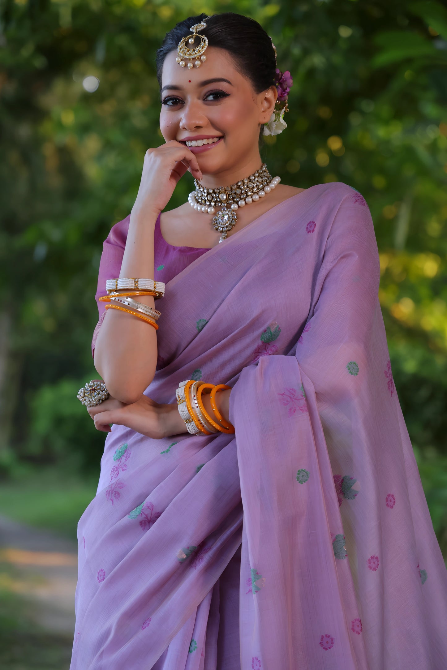 Woman wearing a lavender silk saree with delicate floral embroidery and a matching blouse, accessorized with traditional jewelry and bangles, standing and smiling on a sunlit garden path surrounded by lush green trees and foliage.