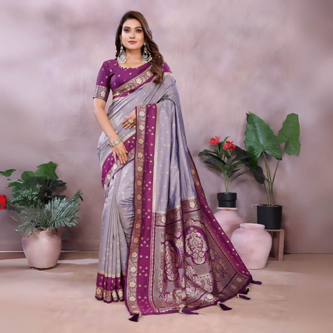 Woman wearing a lavender and purple Banarasi silk saree with an ornate golden border and intricate woven pallu featuring traditional motifs. She pairs the saree with a matching purple blouse adorned with gold detailing and statement jewelry, posing gracefully in a modern studio setting with potted green plants and blush-toned vases in the background.