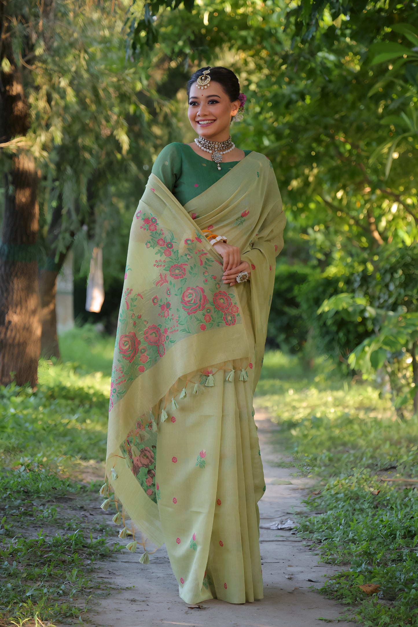 Woman wearing a light yellow silk saree with pastel floral embroidery and a green blouse, accessorized with traditional jewelry and bangles, standing on a sunlit garden path surrounded by lush green trees and foliage