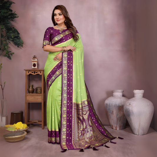 Woman wearing a lime green Banarasi silk saree with a rich purple and gold embroidered border, featuring intricate traditional motifs and an ornate pallu design. She pairs the saree with a matching purple blouse adorned with gold detailing, accessorized with traditional jewelry and bangles, posing gracefully in a modern studio setting with large white vases, a wooden side table, brass decor, green foliage, and a bowl of yellow flowers.