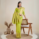 Woman wearing a lime green silk saree with a matching blouse and gold jewelry, elegantly posing beside a wooden chair in a minimalist studio setting with neutral decor, ceramic vases, and pampas grass.