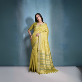 Woman wearing a lime yellow silk saree with subtle stripes and patterned border, paired with a matching blouse and traditional gold jewelry, posing gracefully in a blue-gradient studio backdrop.