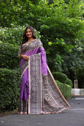 Woman wearing traditional purple Madhubani print saree with intricate designs and ethnic jewelry in a green outdoor setting