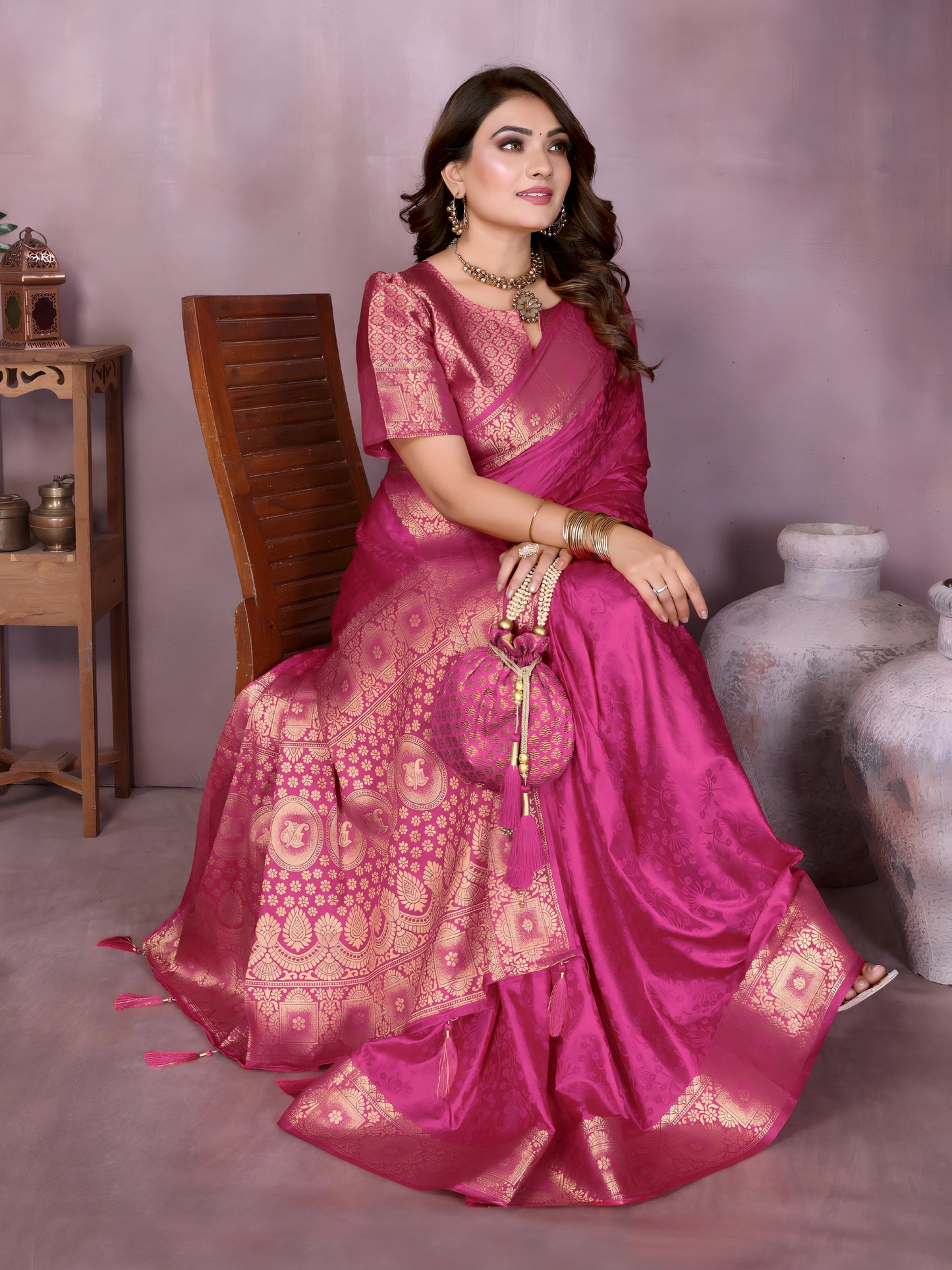 Woman wearing a rich magenta Banarasi silk saree with an ornate golden zari border and intricate woven motifs, paired with a matching short-sleeve blouse and traditional gold jewelry. She poses gracefully in a contemporary studio featuring large white pottery vases, a wooden stand with brass decorative items, a bowl of fresh marigolds, and green foliage, all set against a muted backdrop.