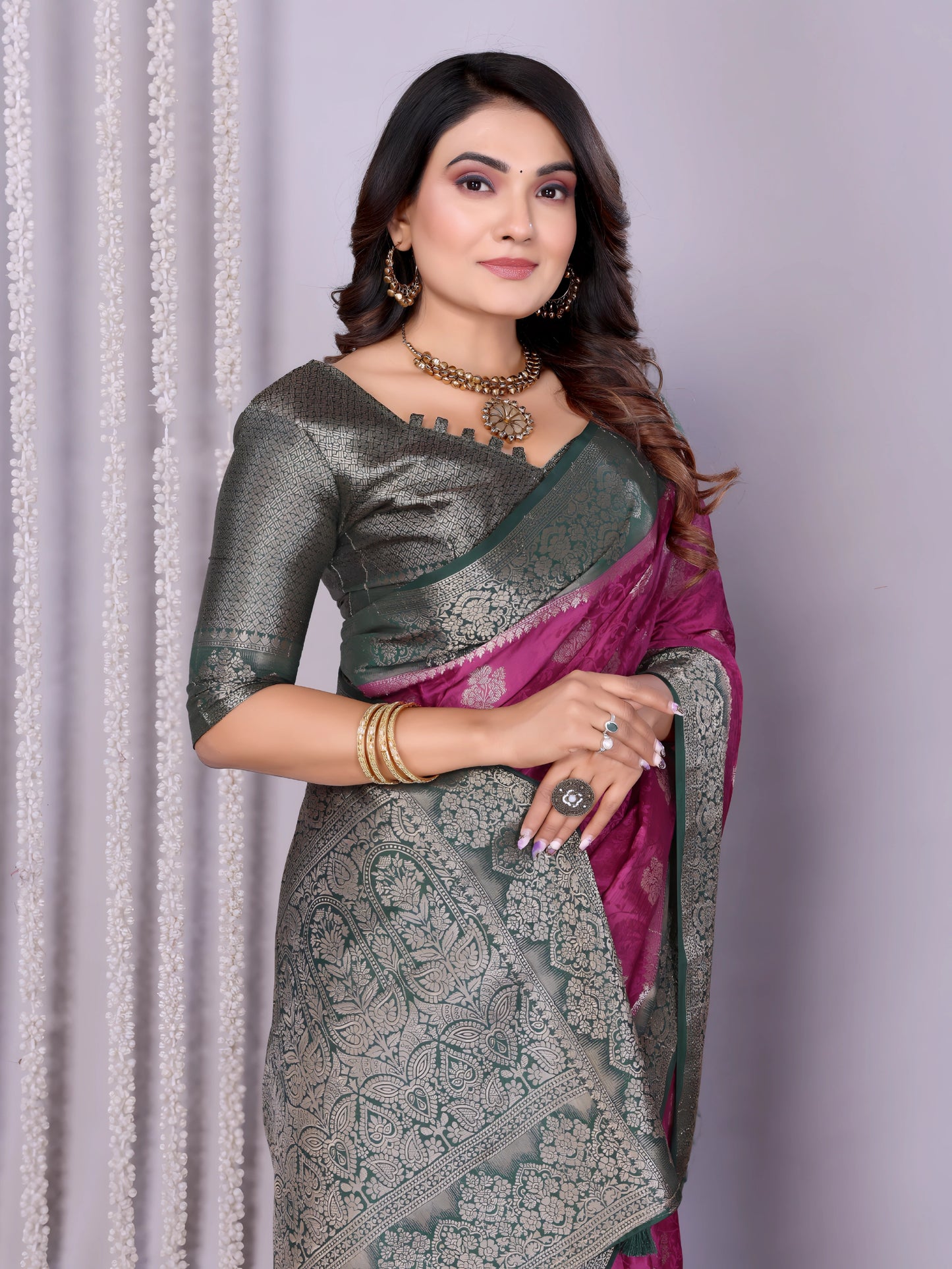 Woman wearing a magenta Banarasi silk saree with a silver and grey zari border, intricate woven motifs, and a matching grey blouse. She is adorned with traditional jewelry and poses gracefully in a modern studio setting featuring woven baskets filled with rose petals and white curtain drapes against a neutral backdrop.
