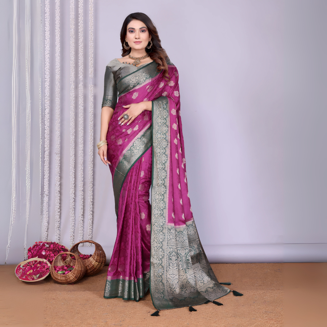 Woman wearing a magenta Banarasi silk saree with a silver and grey zari border, intricate woven motifs, and a matching grey blouse. She is adorned with traditional jewelry and poses gracefully in a modern studio setting featuring woven baskets filled with rose petals and white curtain drapes against a neutral backdrop.