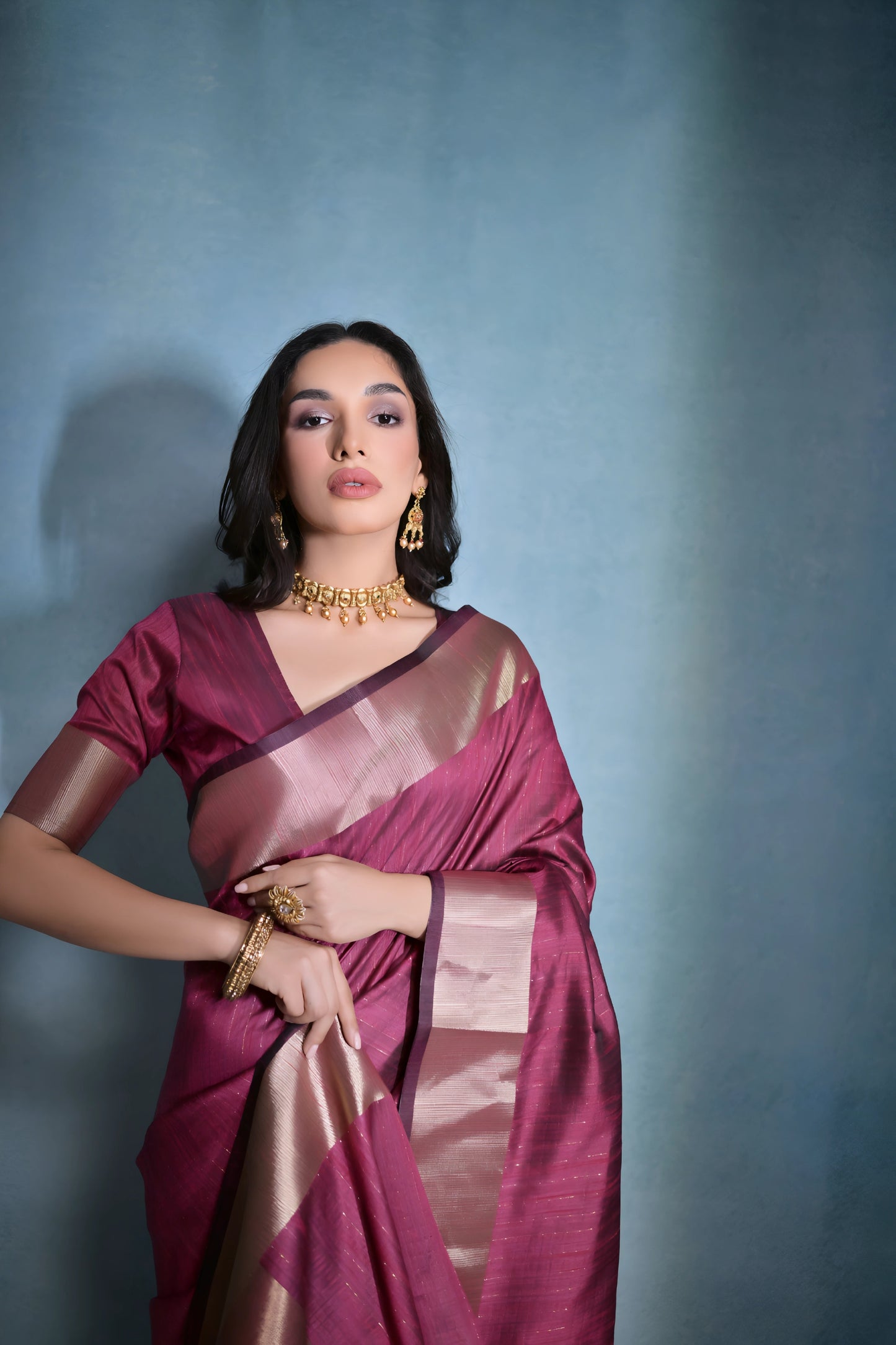 Woman wearing a magenta silk saree with a gold border and traditional jewelry, standing against a blue background.