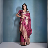 Woman wearing a magenta silk saree with a gold border and traditional jewelry, standing against a blue background.