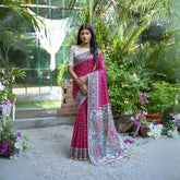 Woman wearing a magenta saree with intricate white tribal-inspired prints and a multicolor pallu featuring circular and geometric patterns, paired with a short-sleeve embroidered blouse and traditional jewelry, standing gracefully in a lush outdoor garden surrounded by tropical plants and blooming flowers.