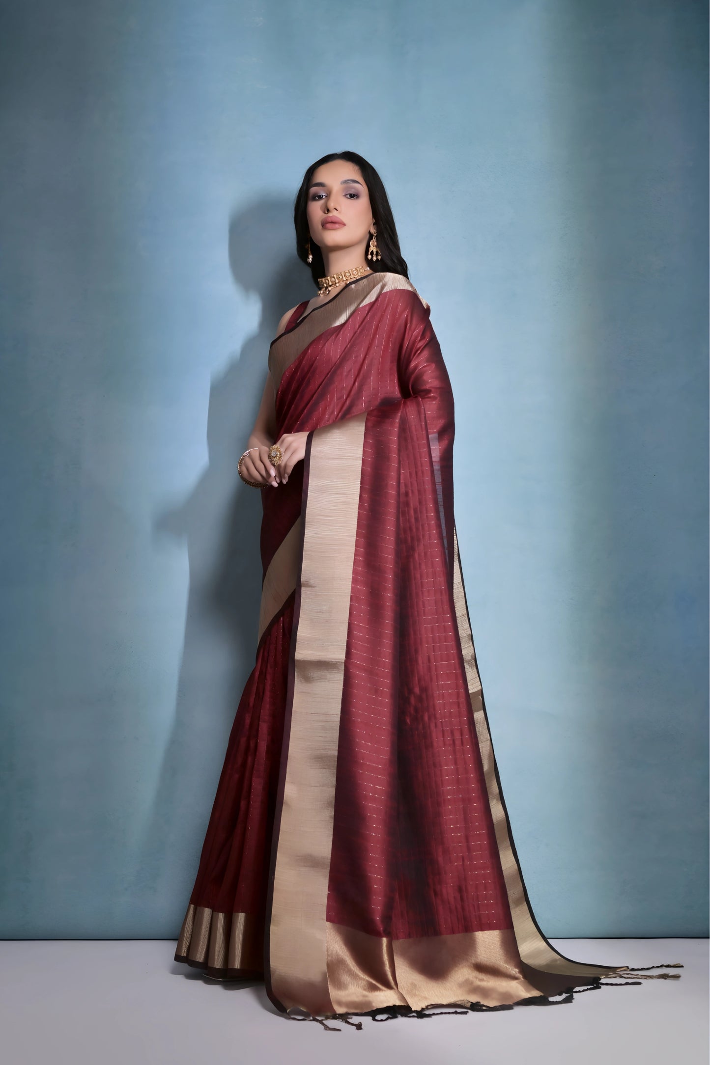 Woman wearing a maroon silk saree with gold border and traditional jewelry, standing against a blue background.