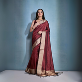 Woman wearing a maroon silk saree with gold border and traditional jewelry, standing against a blue background.