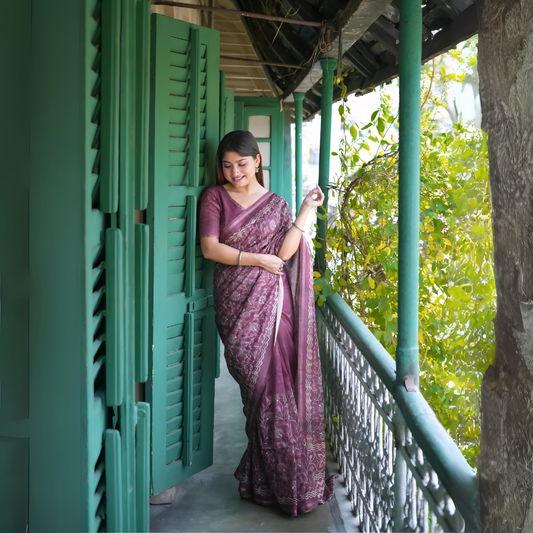 Woman wearing a mauve floral printed saree with a matching blouse, standing on a vintage balcony with green shutters and railings, surrounded by leafy vines and natural light.