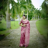 Woman wearing a mauve handloom cotton saree with white tribal-inspired prints and matching blouse, accessorized with traditional jewelry, standing on a stone pathway surrounded by lush green palm trees in a garden setting.