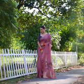 Woman wearing a mauve silk saree with subtle floral embroidery and a navy blue blouse, accessorized with traditional jewelry and red bangles, standing beside a white picket fence in a sunlit garden surrounded by lush green trees.