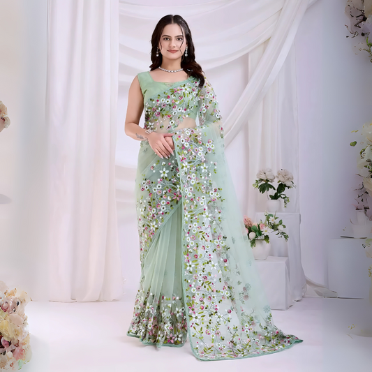 Woman wearing a mint green net saree with intricate multicolor floral embroidery and a matching sleeveless blouse, posing in a modern studio setting with white draped curtains and floral arrangements.