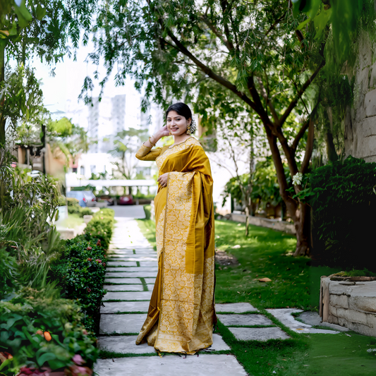 Woman wearing a mustard yellow brocade silk saree with intricate gold floral patterns and a matching blouse, standing gracefully on a stone pathway in a lush green garden with manicured lawns, trees, and vibrant foliage in the background.