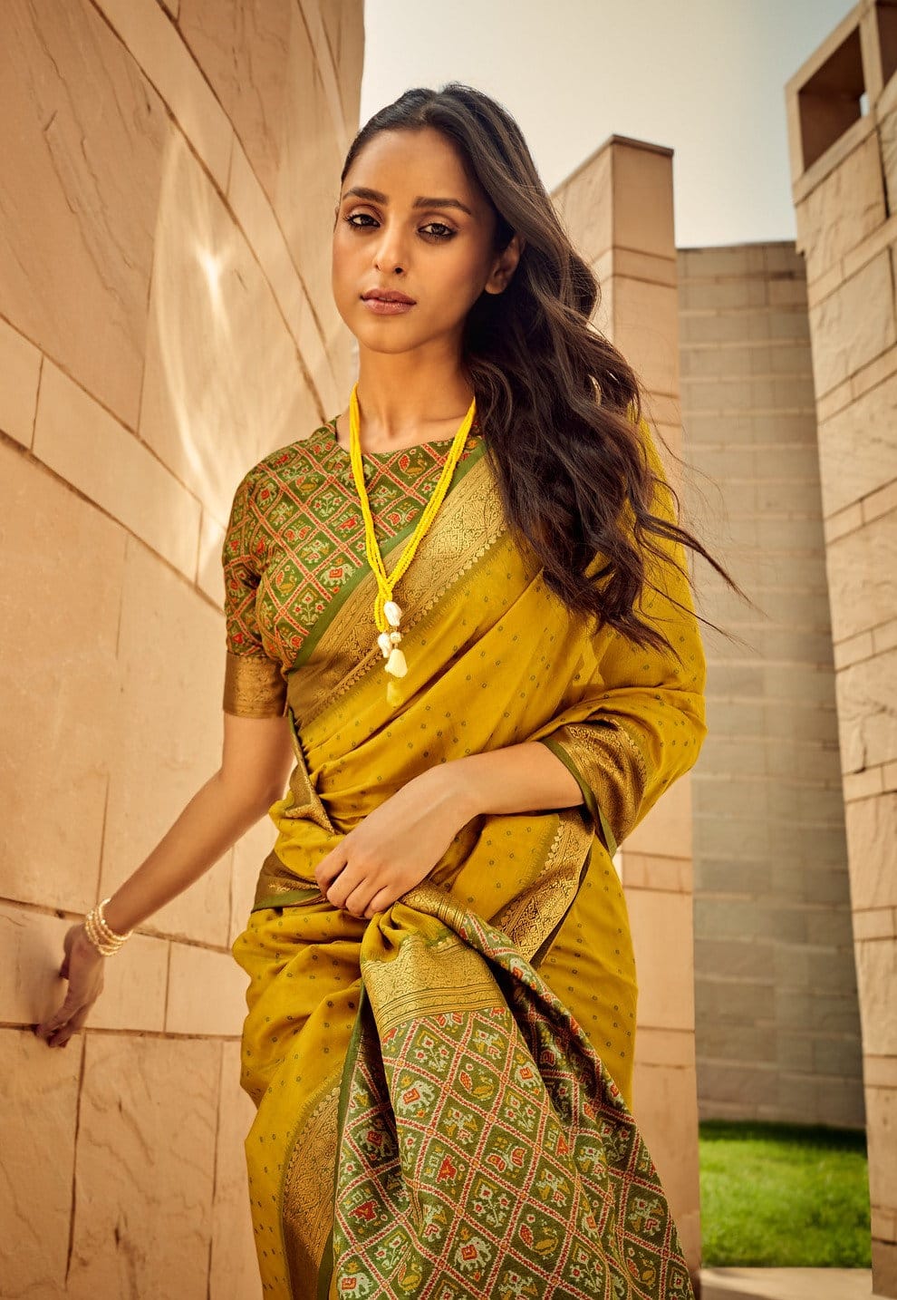 Woman wearing a mustard yellow saree with intricate zari work and green embroidered blouse, exuding traditional Indian elegance in a modern architectural setting