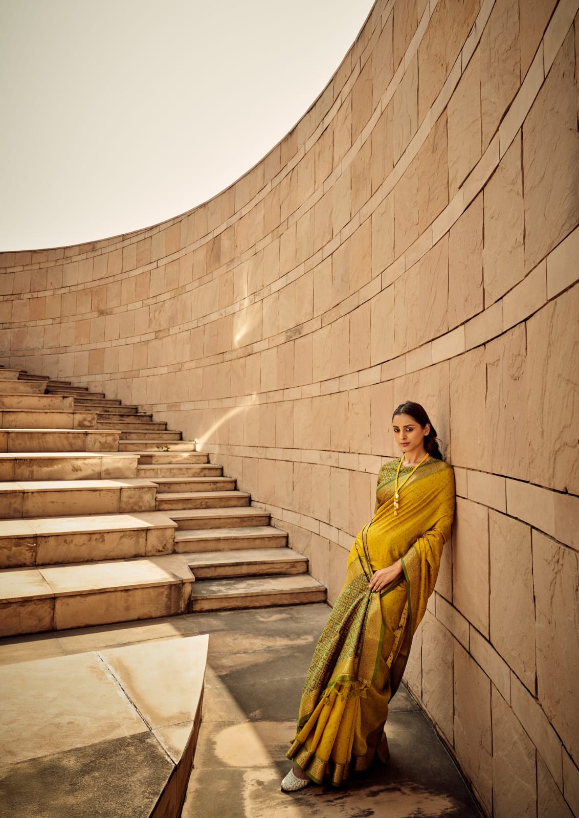 Woman wearing mustard yellow saree with intricate zari work, standing against a curved stone wall with stairs in the background, showcasing modern ethnic fashion elegance