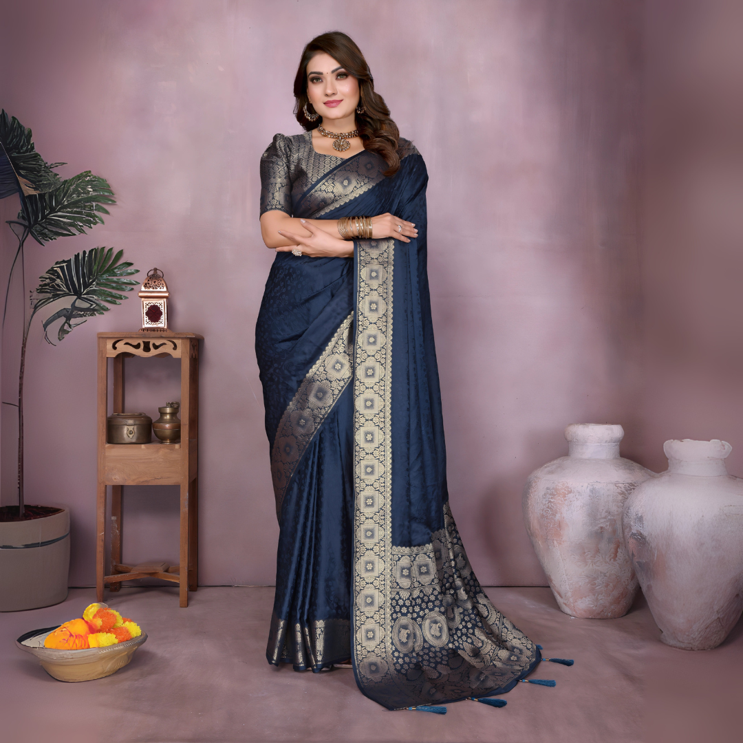 Woman wearing a navy blue Banarasi silk saree featuring an ornate golden zari border with traditional circular woven motifs, paired with a matching metallic blouse and gold jewelry. She poses elegantly in a modern studio decorated with large rustic white clay pots, a wooden stand displaying brass decorative items, a bowl of fresh marigold flowers, and green potted foliage, all set against a soft mauve backdrop.