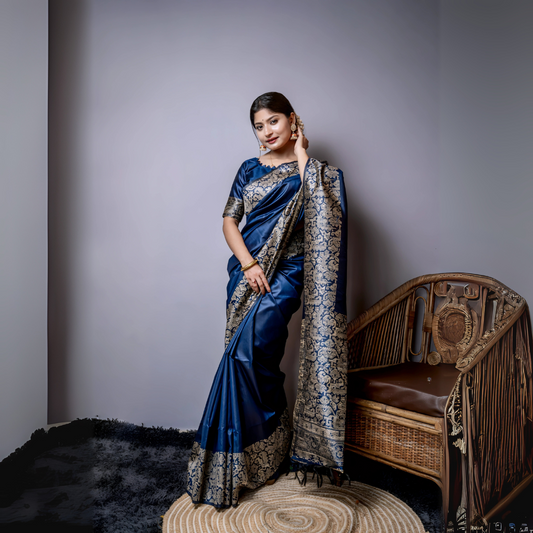 Woman wearing a navy blue silk saree with intricate silver brocade border and pallu, paired with a matching blouse and gold bangles, standing gracefully beside a vintage wooden chair in a minimalist modern studio with grey walls and a textured rug.