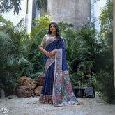 Woman wearing a navy blue saree with intricate white tribal-inspired prints and a multicolor pallu featuring circular and geometric patterns, paired with a beige embroidered blouse and traditional jewelry, standing gracefully in a lush outdoor garden surrounded by tropical plants.