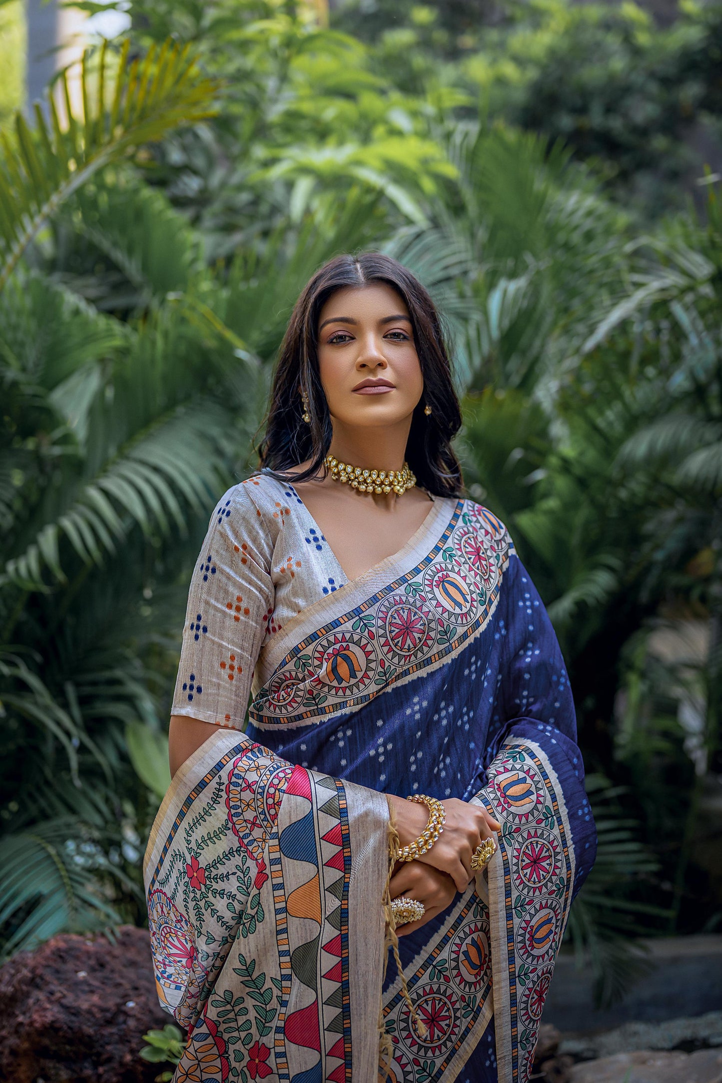 Woman wearing a navy blue saree with intricate white tribal-inspired prints and a multicolor pallu featuring circular and geometric patterns, paired with a beige embroidered blouse and traditional jewelry, standing gracefully in a lush outdoor garden surrounded by tropical plants.