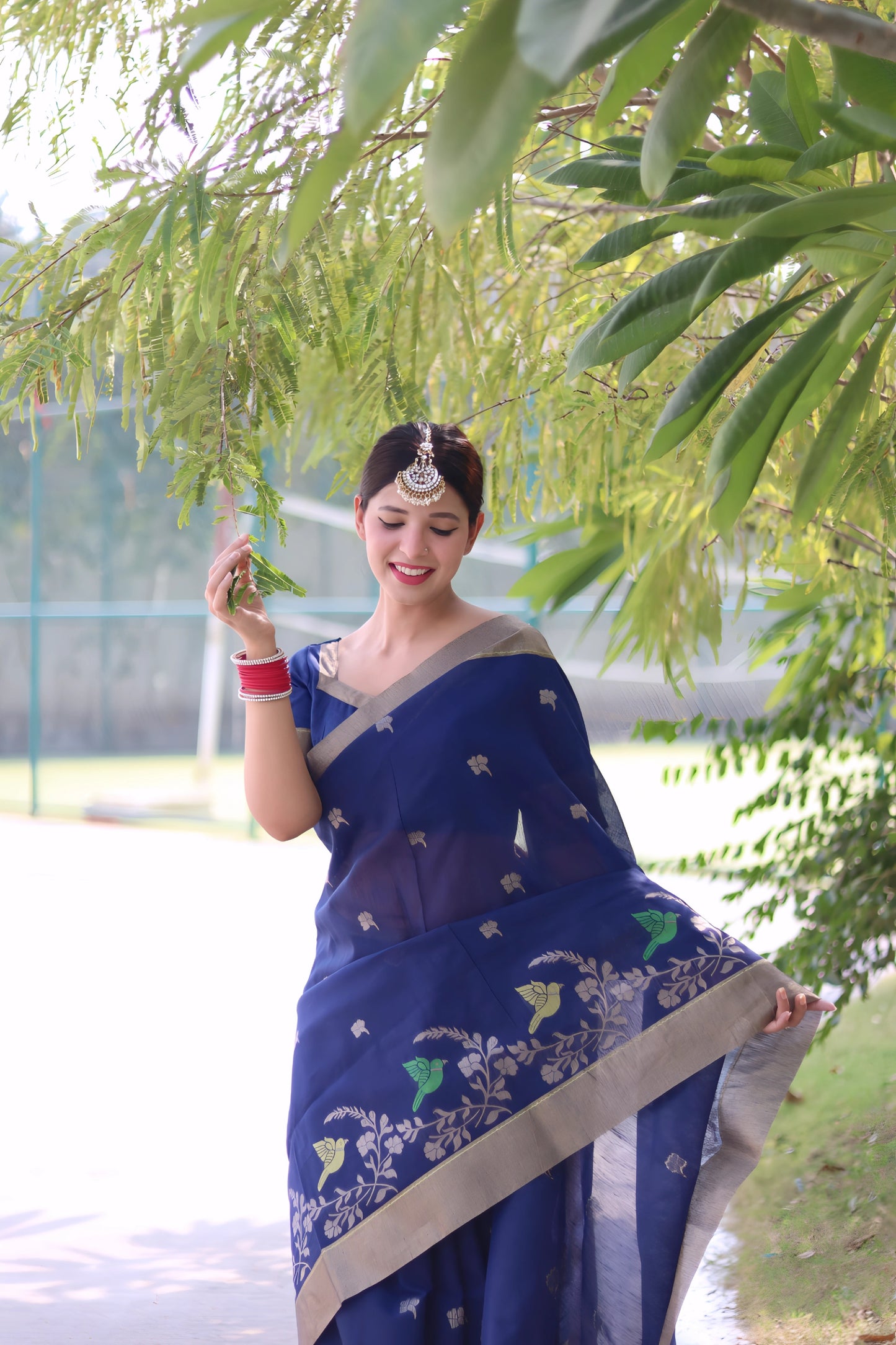 Woman wearing a navy blue saree with gold border and floral embroidery, posing outdoors under leafy trees on a sunlit pathway.