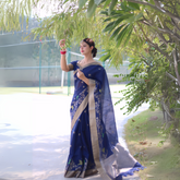 Woman wearing a navy blue saree with gold border and floral embroidery, posing outdoors under leafy trees on a sunlit pathway.