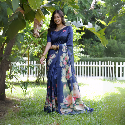 Woman wearing a navy blue saree with large handpainted lotus and leaf floral motifs, accessorized with traditional gold jewelry and bangles, standing on a lush green lawn beneath tropical trees with a white picket fence in the background