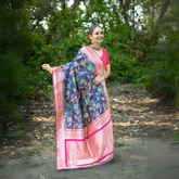 Woman wearing a navy blue silk saree with vibrant multicolor floral motifs and a contrasting pink border, paired with a pink blouse and traditional jewelry, standing gracefully on a shaded garden path surrounded by lush greenery and trees.