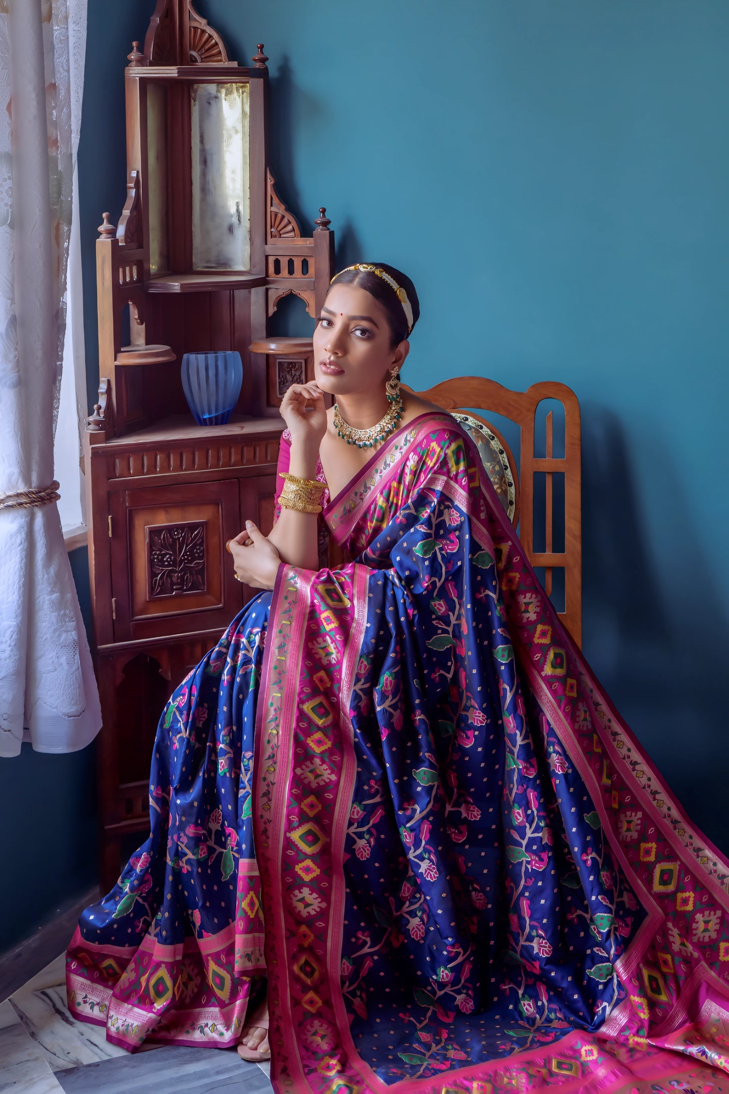 Woman wearing a navy blue and pink Patola silk saree with intricate geometric and floral patterns, paired with a matching blouse and traditional jewelry, elegantly posing beside a vintage wooden chair in a classic bedroom setting with teal blue walls, wooden furniture, and decorative accents.