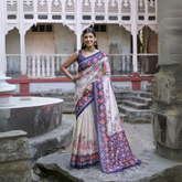 Woman wearing an off-white silk saree with blue and red Patola-inspired geometric and floral patterns, paired with a matching blue blouse and traditional jewelry, elegantly posing in a vintage courtyard with stone steps, ornate pillars, and a heritage building in the background.