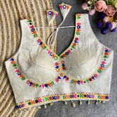 Off-white blouse featuring vibrant mirror work and traditional Kutch multicolor embroidery along the neckline and hem, with delicate sequin accents and shell tassels. The blouse includes a stylish back tie-up with matching embroidered tassels and is displayed flat on a textured surface with a floral bouquet, highlighting its festive ethnic elegance.