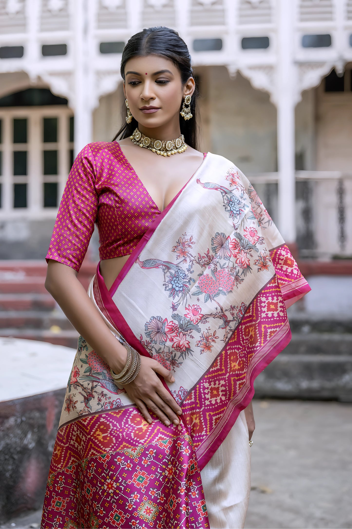 Woman wearing an off-white silk saree with vibrant pink Patola-inspired geometric and floral borders, featuring intricate floral motifs, paired with a matching pink blouse and traditional jewelry, elegantly posing on stone steps in front of a vintage heritage house with ornate railings and classic architectural details.