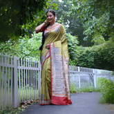 Woman wearing an olive green Madhubani-printed saree with a vibrant red border and intricate traditional motifs, paired with a matching blouse and long braid, elegantly standing on a garden path lined with a white fence and surrounded by lush green foliage.