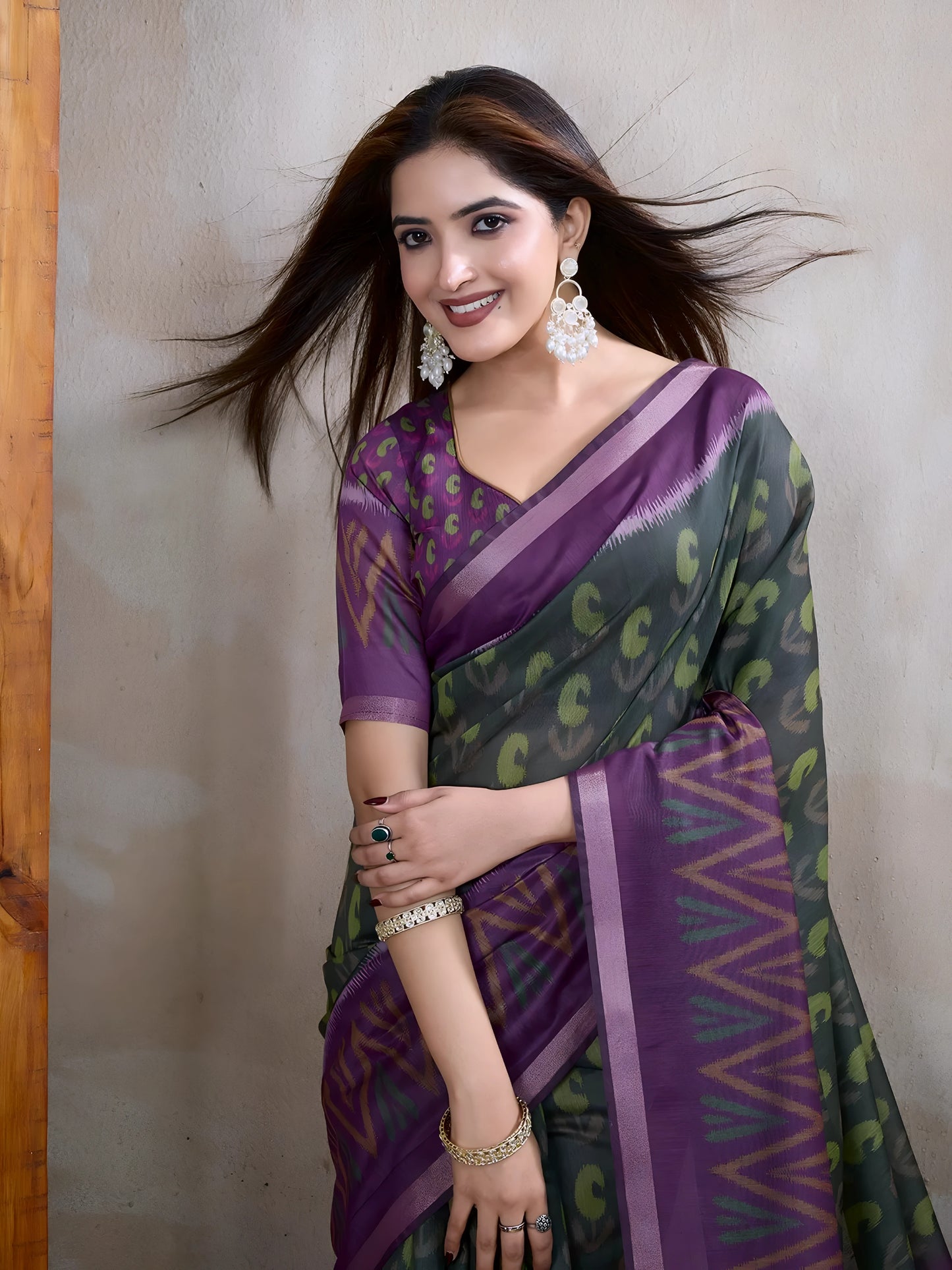 Woman wearing an olive green saree adorned with purple geometric patterns and bold abstract motifs, paired with a matching blouse and statement silver earrings. She poses elegantly in a contemporary studio setting against a neutral textured wall, with a tall woven screen and rustic wooden flooring, showcasing the saree’s vibrant pallu and intricate design.